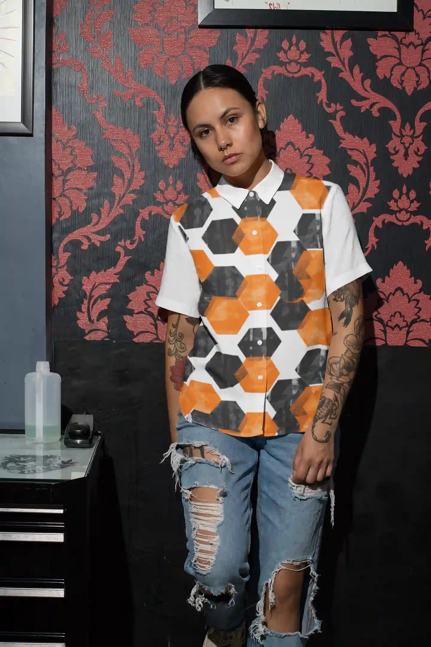 Person wearing a patterned shirt and ripped jeans against a decorative wall.