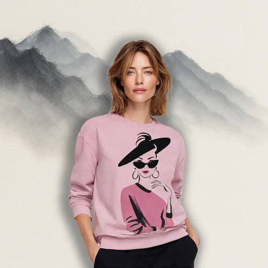 A woman with short brown hair wearing the pink 'City Glamour Muse' sweatshirt with black pants against a painted mountain backdrop, showcasing its stylish unisex fit.