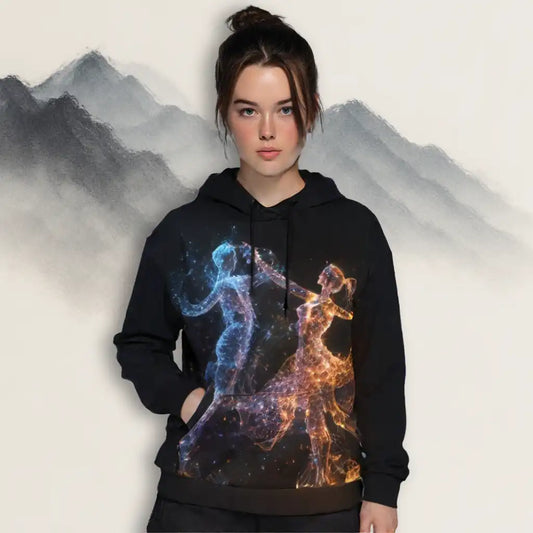 Female model wearing a black hoodie with a vibrant particle art print featuring dancing figures in blue and orange light, standing against an artistic mountain backdrop. Shows the wearable art hoodie's fit and lifestyle context.