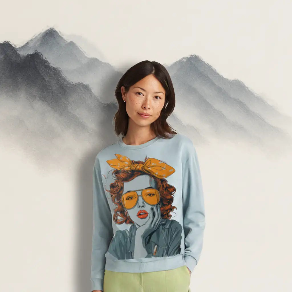 A woman with dark hair and freckles wearing the light blue 'Retro Pop Muse' sweatshirt with light green pants against a painted mountain backdrop, showcasing its fit.