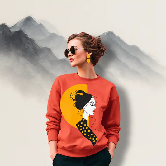 A woman with wavy brown hair wearing the vibrant orange 'Sunset Muse' sweatshirt with white pants in a modern room, showcasing its stylish fit.